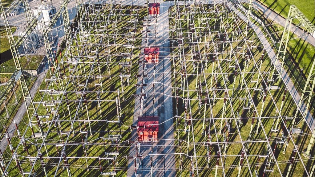 Aerial view of an electrical substation with a grid of metal structures, high‑voltage lines, and green support poles. Several small red‑roofed equipment buildings line a central paved path.